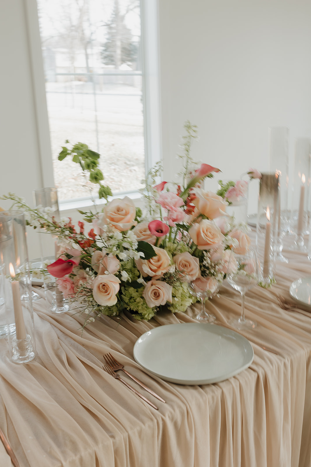Soft pastel wedding tablescape with blush linens, peach and pink floral centerpiece, taper candles, and glassware near a window, styled using a wedding vendor checklist for cohesive reception décor.