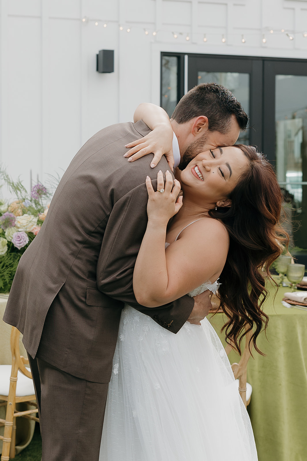 Bride and groom hugging and laughing during an outdoor editorial wedding portrait in front of a modern white venue with green tables and string lights, inspired by a wedding vendor checklist for styling.