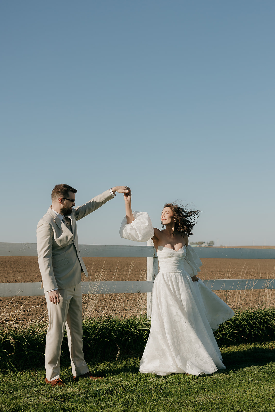 Bride and groom twirling and dancing in a field by a white fence during golden hour, capturing a joyful and cinematic wedding portrait moment.