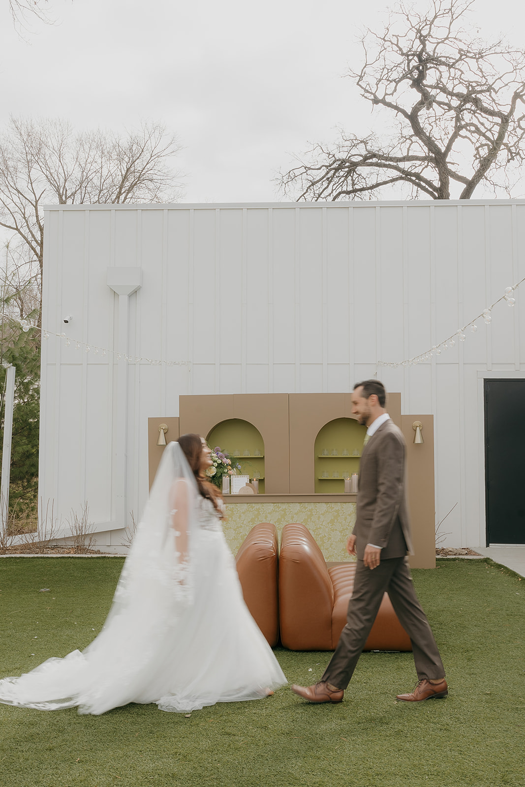 Bride and groom walking toward each other in front of a modern outdoor bar setup with architectural arches and string lights during an editorial wedding shoot.