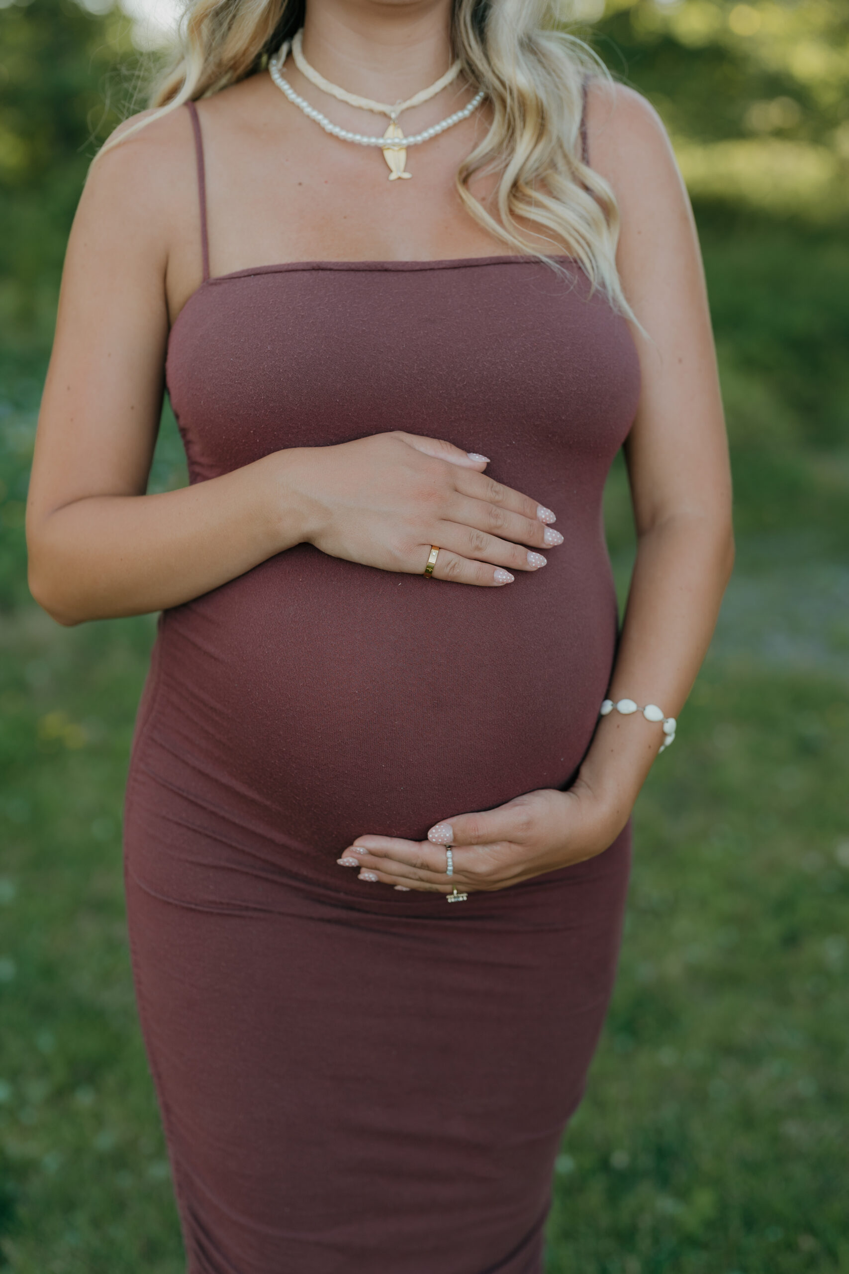 The pregnant wife’s hands rest gently on her baby bump, focusing on detail, emotion, and anticipation in a quiet outdoor setting.