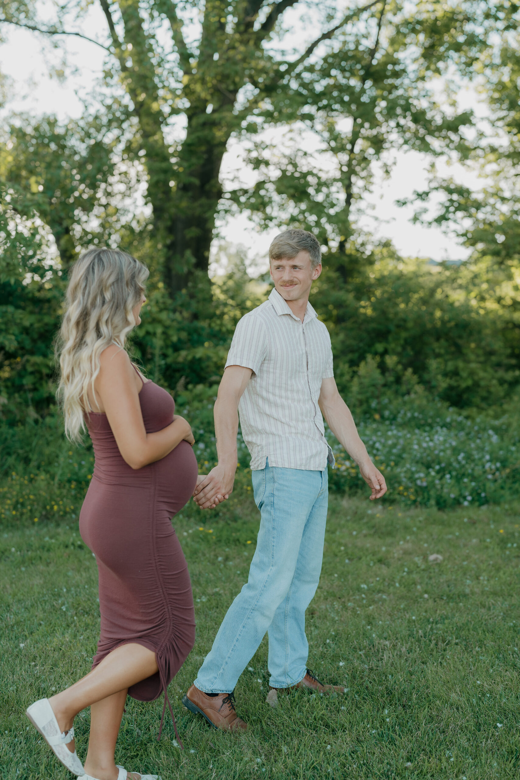 The couple walks together across the grass, the wife’s hand resting on her belly as the husband looks back at her with a smile.