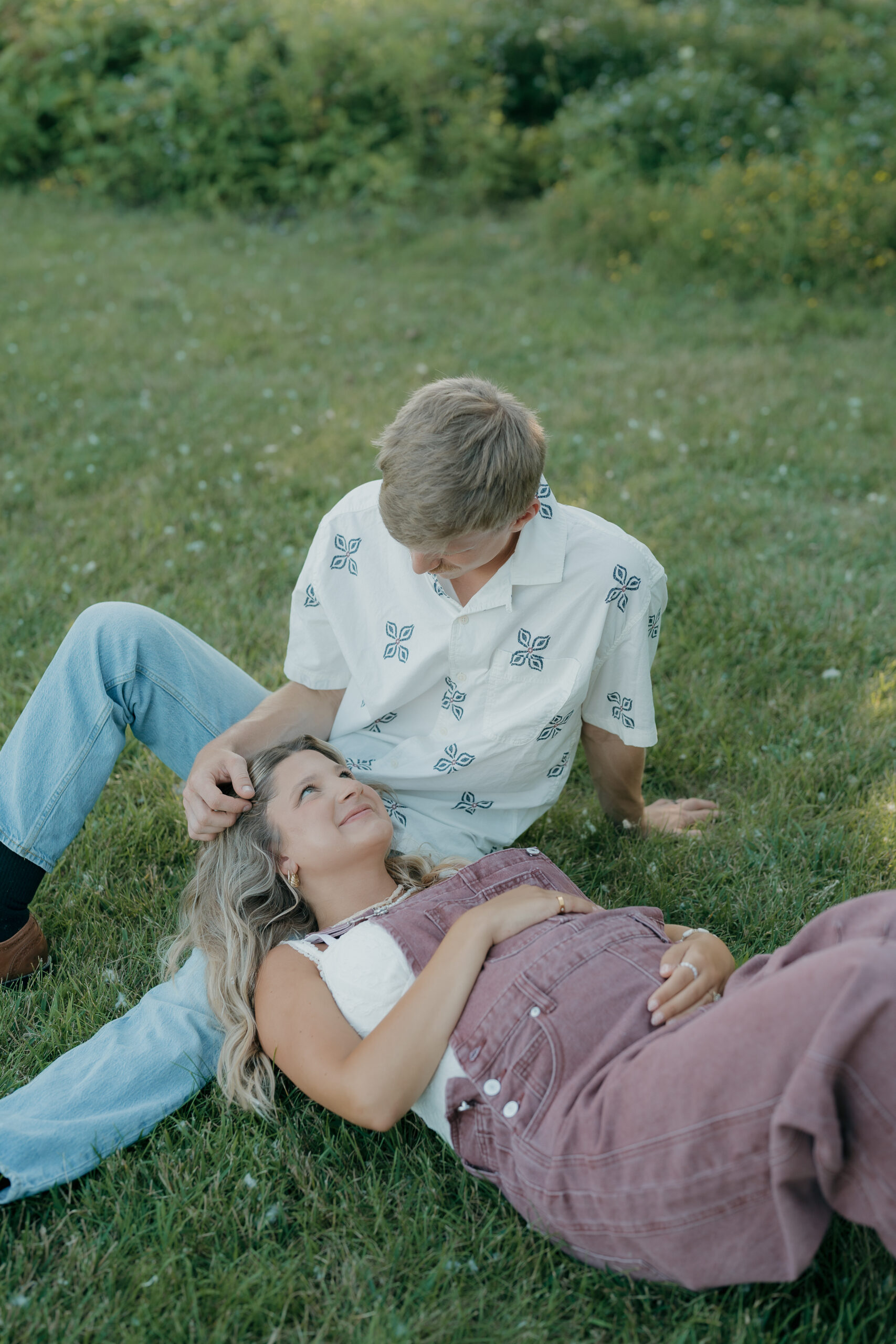 A husband and wife sit together on the grass, relaxed and smiling as they lean into one another surrounded by greenery.