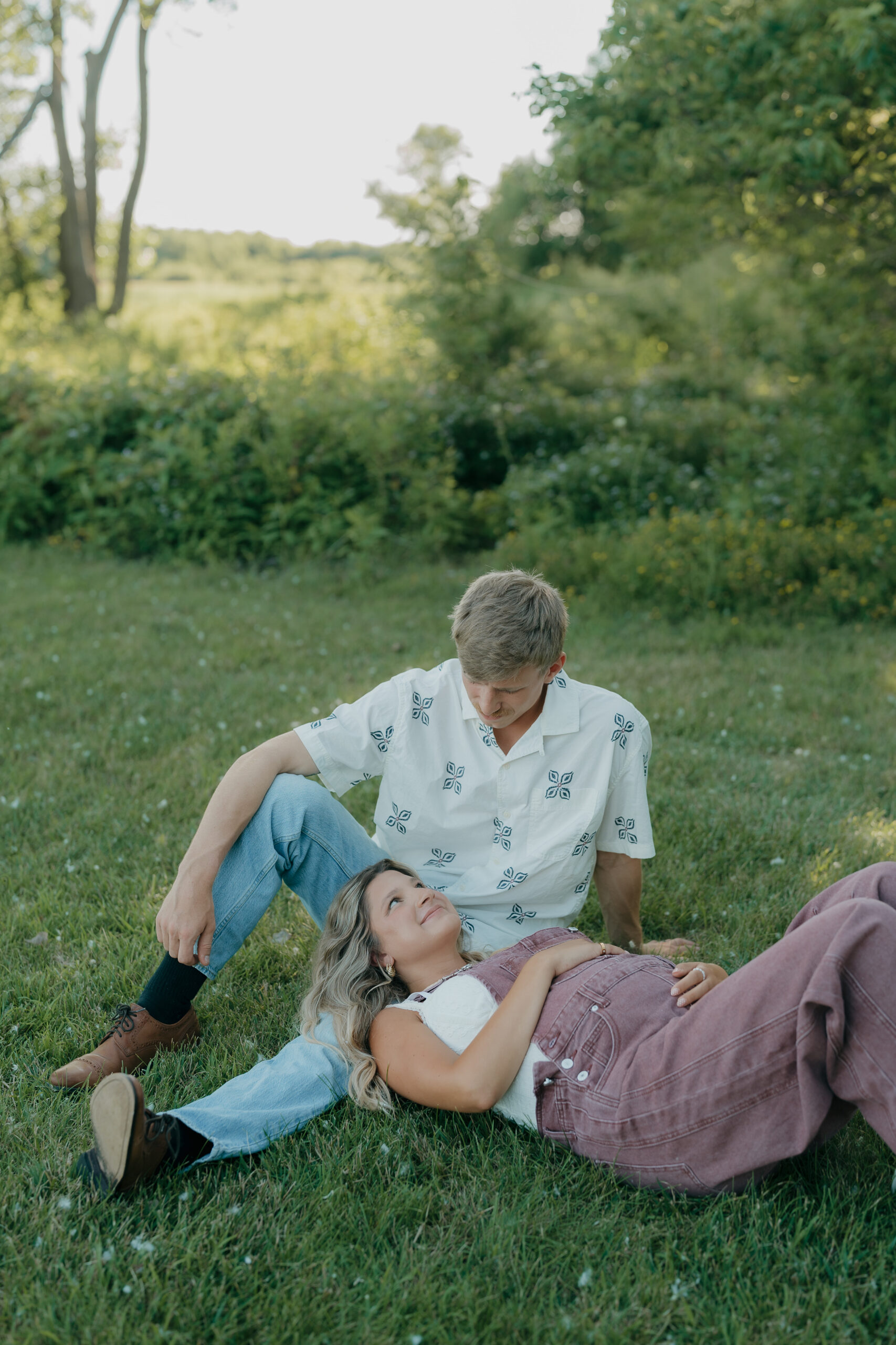 A candid moment of the pregnant soon to be mom lying back on the grass while her husband partner looks down at them, framed with a relaxed documentary wedding photography approach.