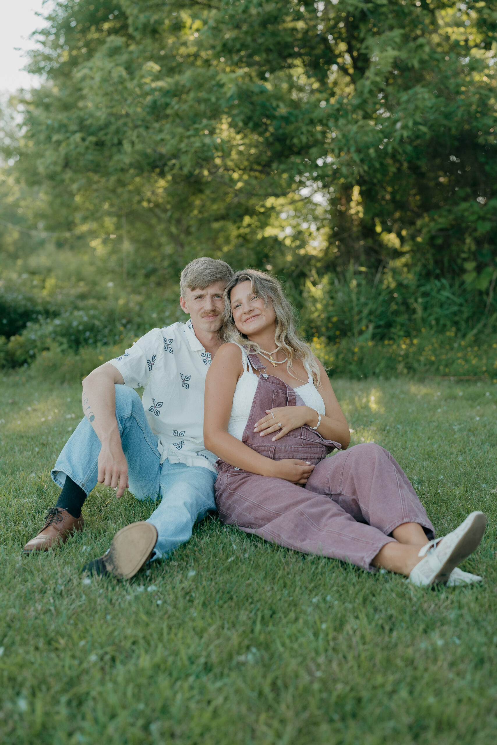 A husband reclines on the grass while his pregnant wife lies beside him, looking up at him with a soft smile, framed through documentary wedding photography.