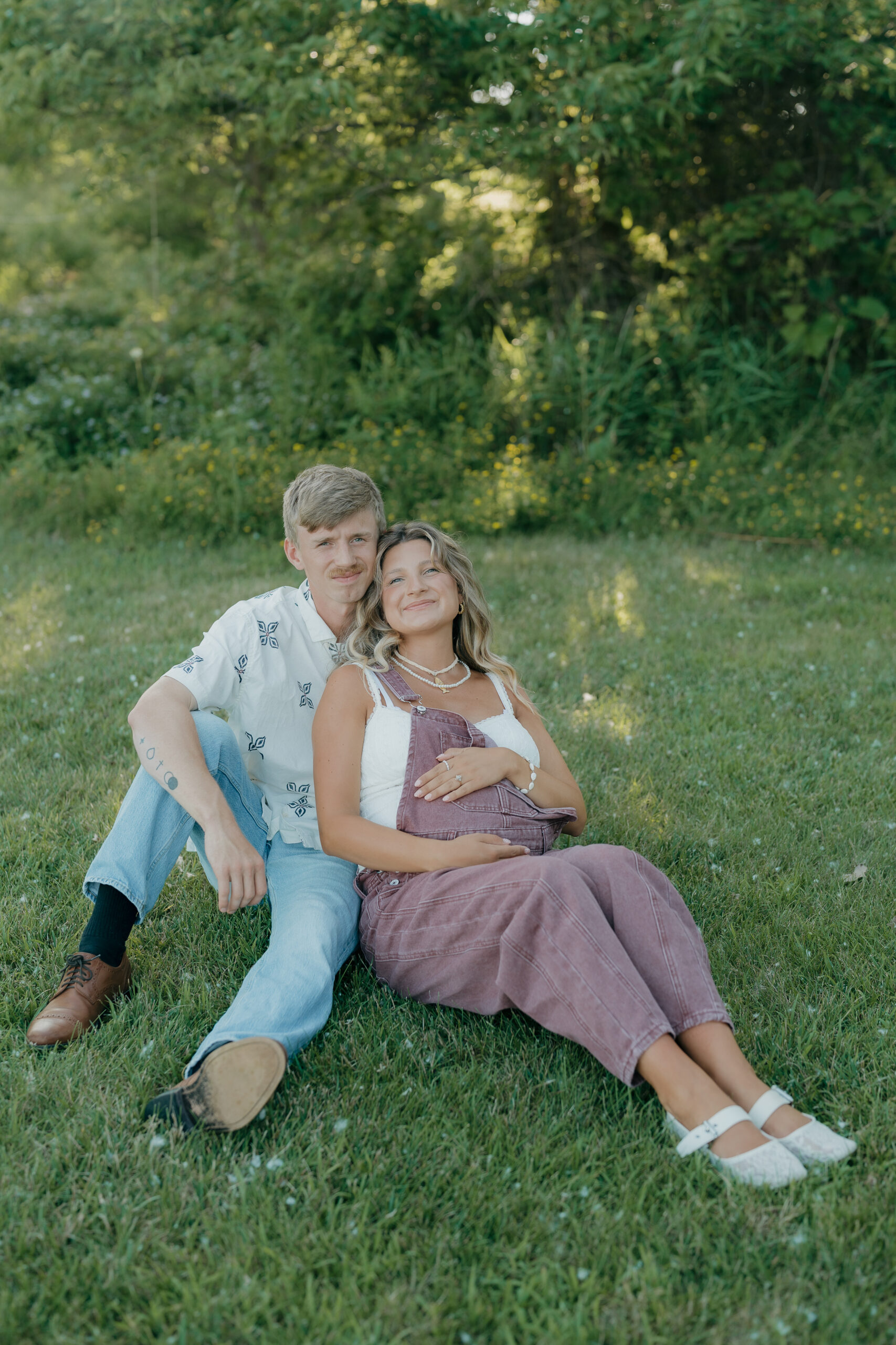 The expecting couple sits together on the grass, smiling softly as they lean into one another surrounded by greenery.