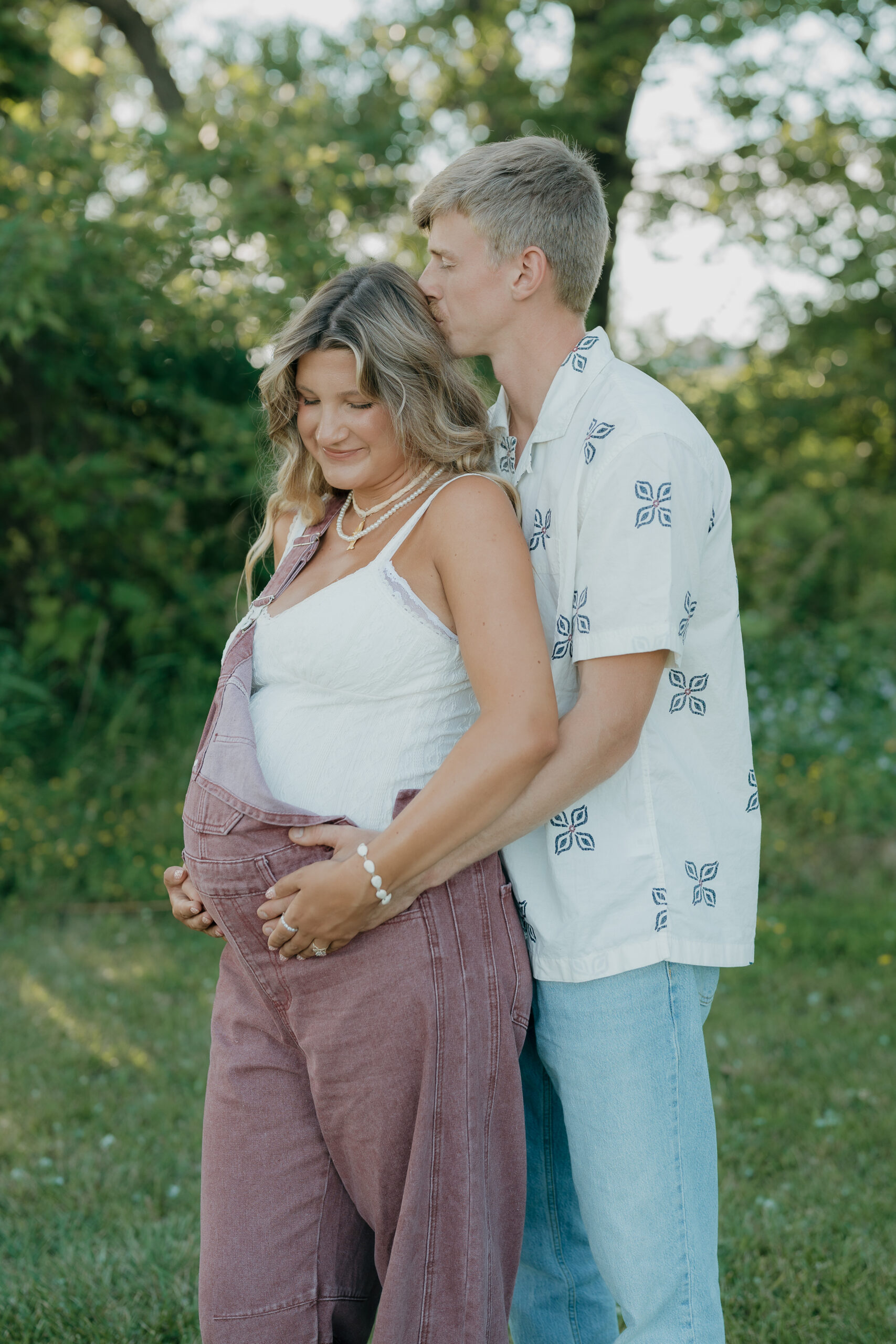 A husband stands behind his pregnant wife in a grassy field, gently kissing the top of her head while holding her baby bump, captured with the emotional storytelling of documentary wedding photography.