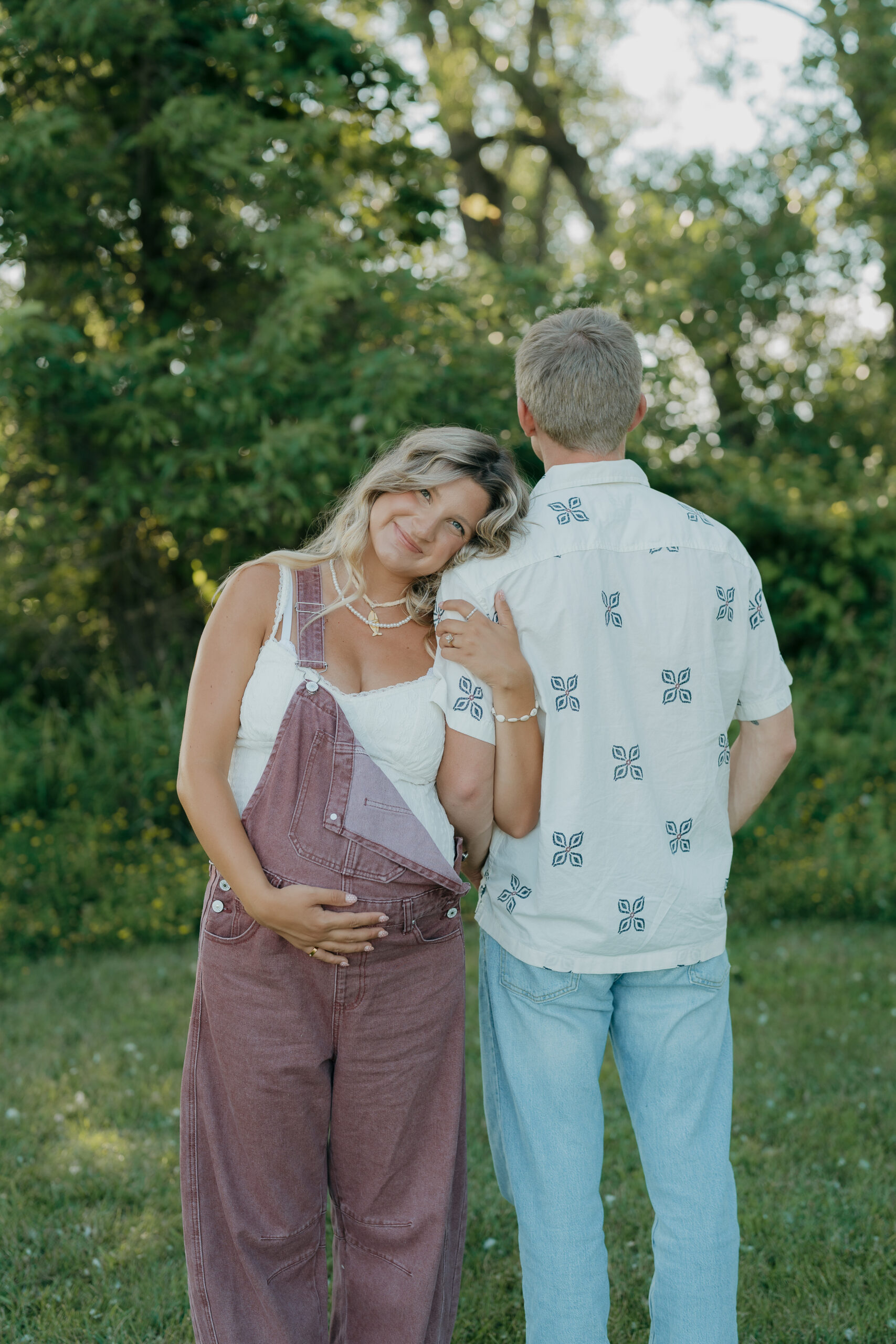 A back-facing portrait of the couple standing together, with the soon to be mom leaning her head on her husbands shoulder in soft evening light.