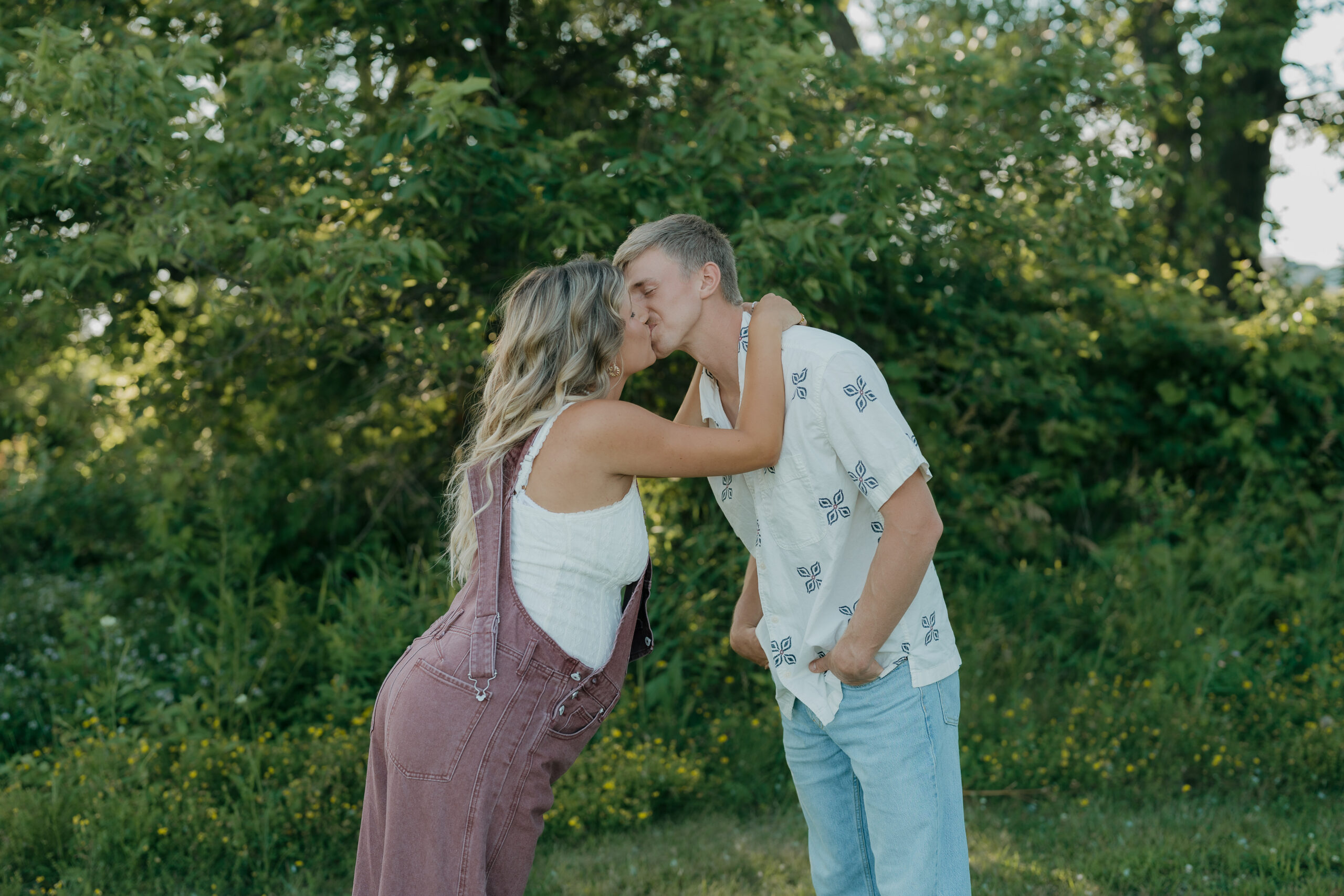 The couple shares a sweet kiss outdoors, surrounded by trees and wildflowers in a natural, unposed moment.