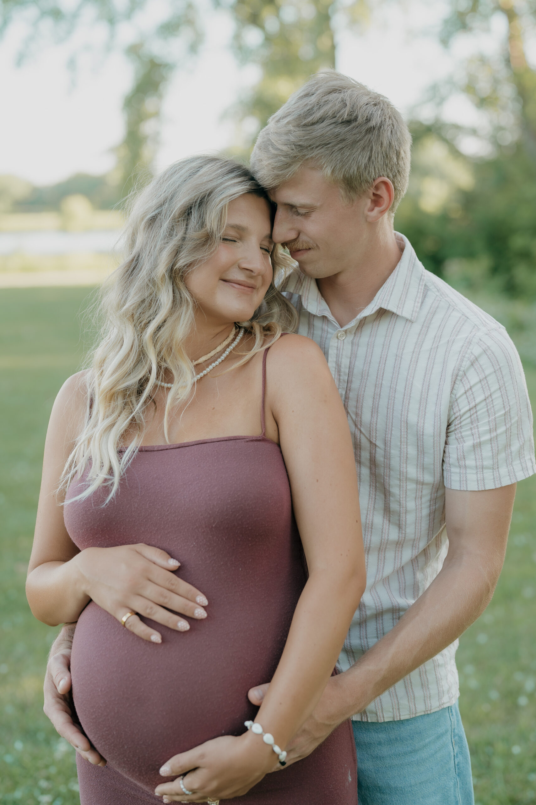 A maternity couple stands close together in a grassy field, the husband's arms wrapped gently around the baby bump, captured with the intimacy of documentary wedding photography.