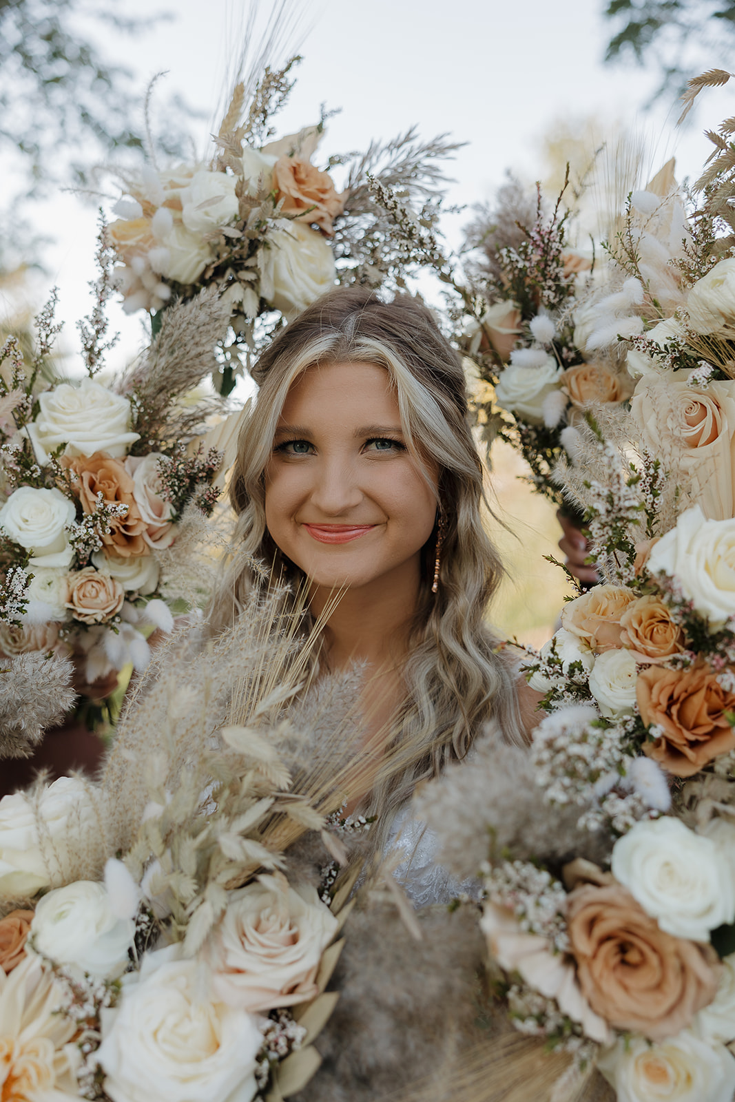 A bride smiles through a floral arch made of neutral-toned roses, dried florals, and textured greenery.