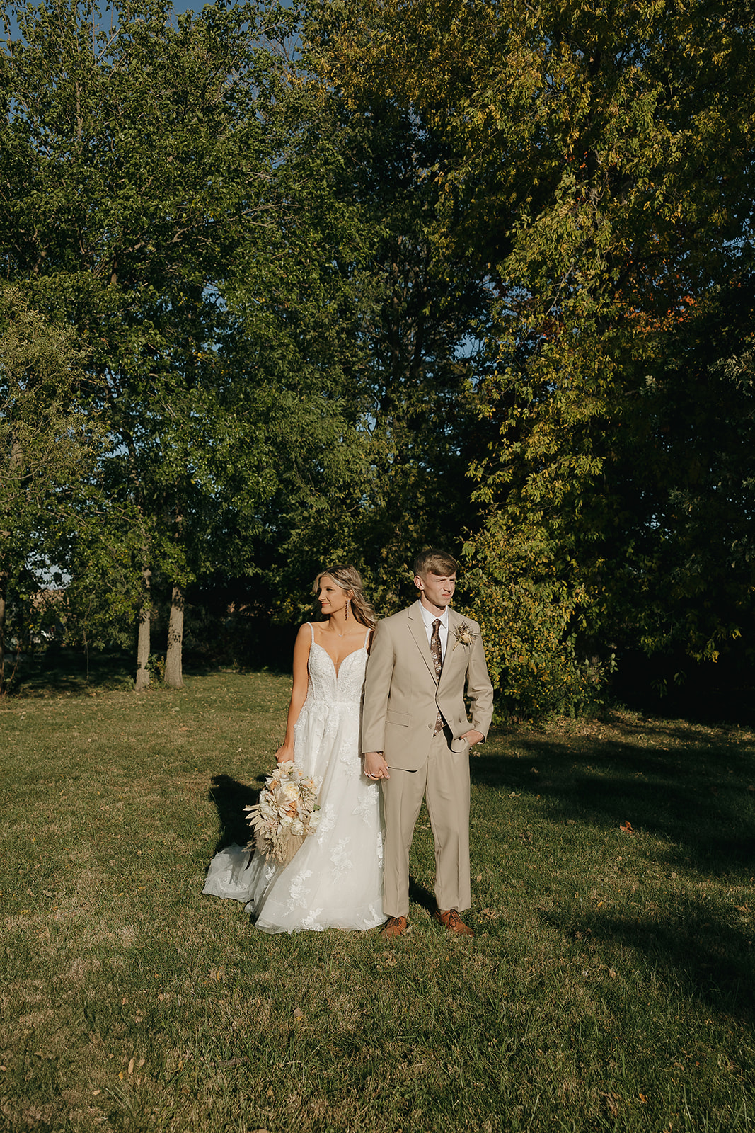 The bride and groom walk hand in hand across an open field, sunlight filtering through the trees as they move naturally together.