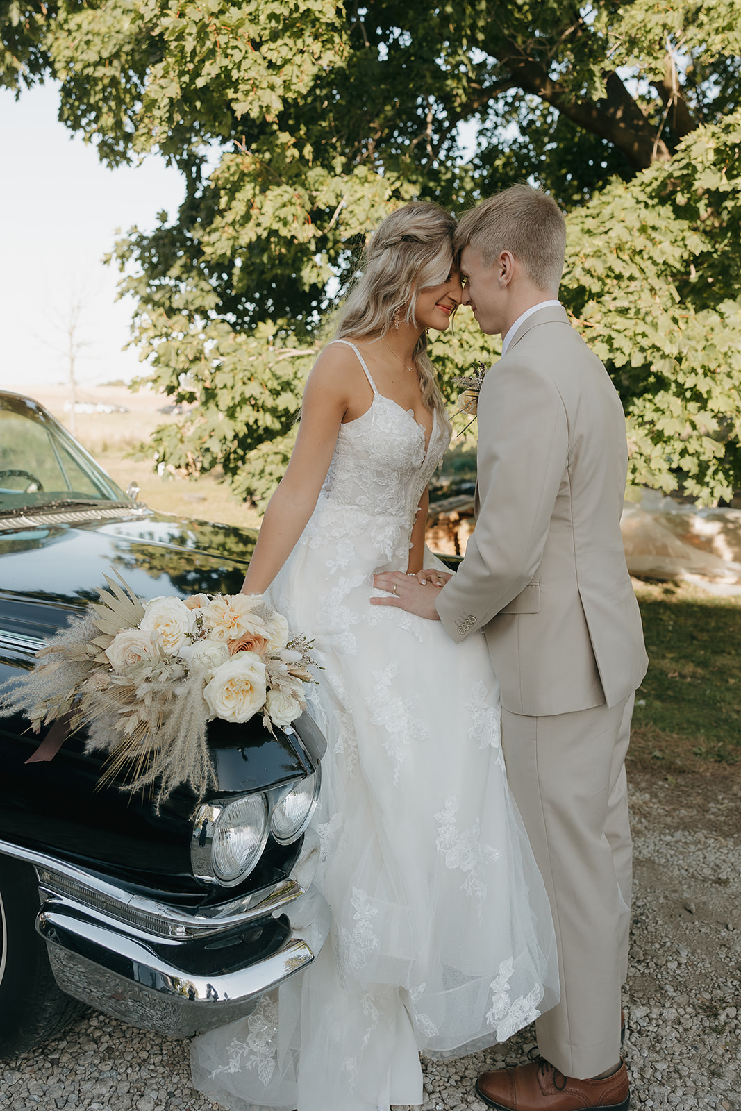 A bride and groom stand close beside a vintage black car, foreheads touching as sunlight filters through the trees, captured with documentary wedding photography.