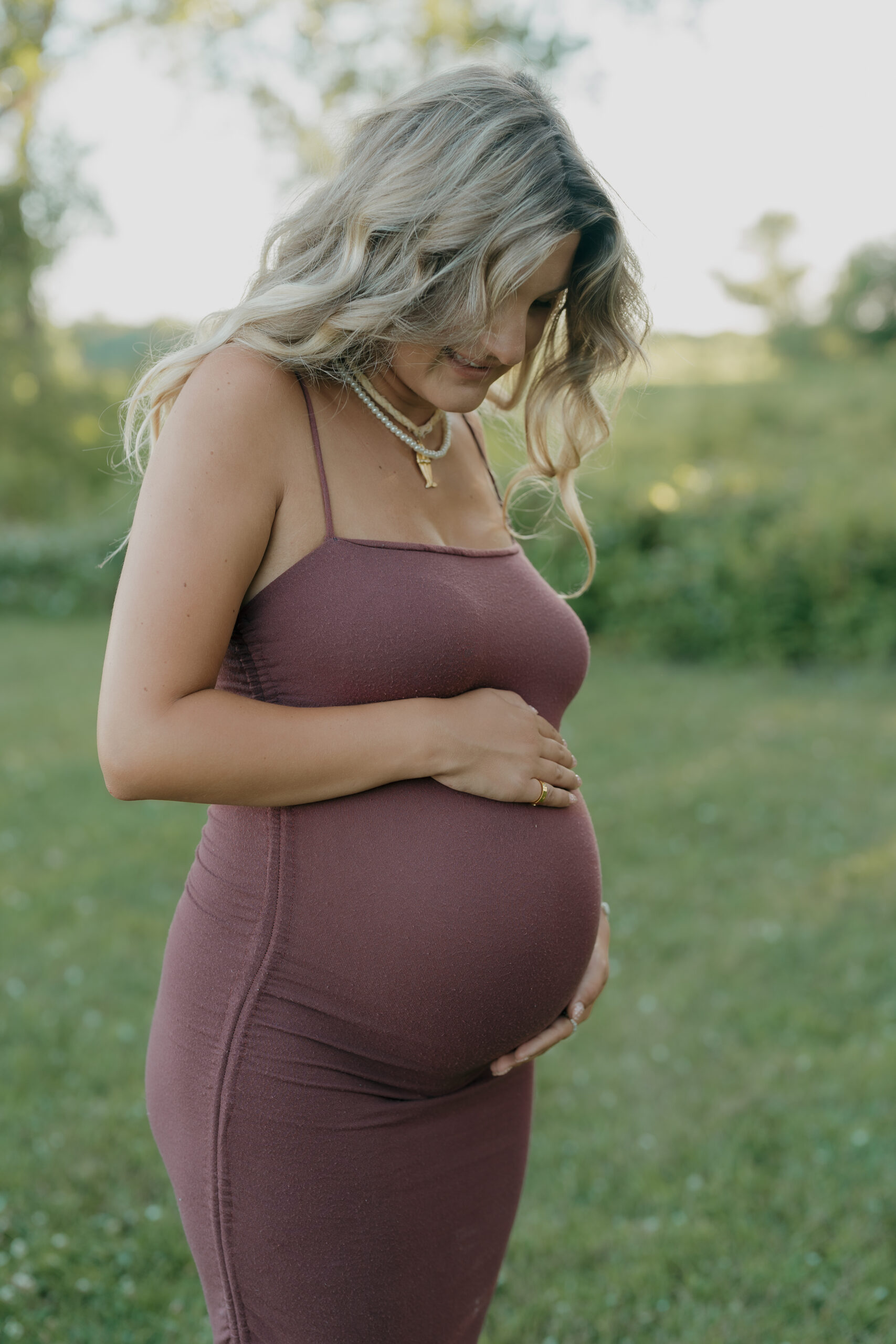 A pregnant woman looks down at her belly, her hair softly framing her face in a peaceful outdoor setting.