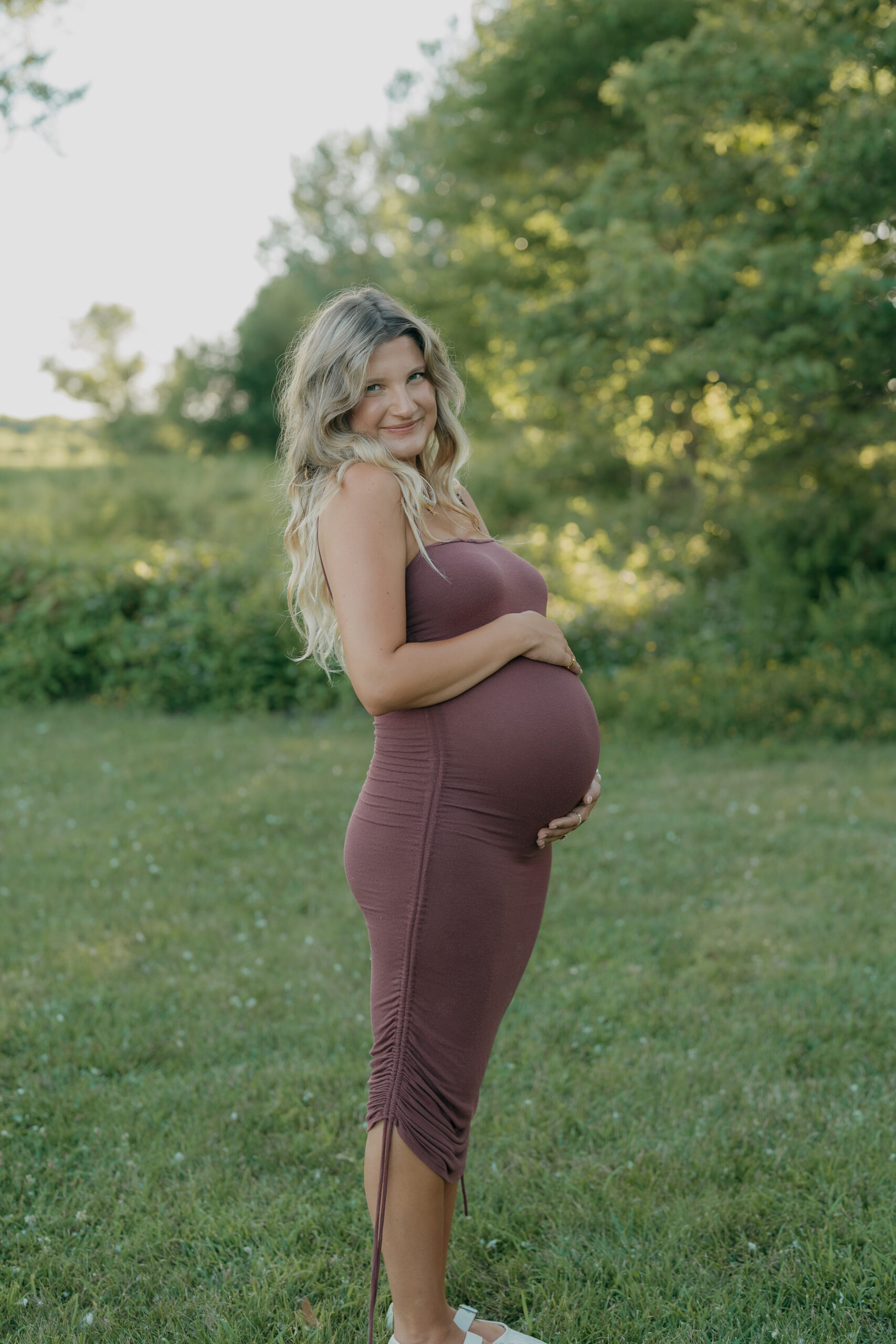 A pregnant woman smiles gently while standing in tall grass, her posture relaxed and confident.