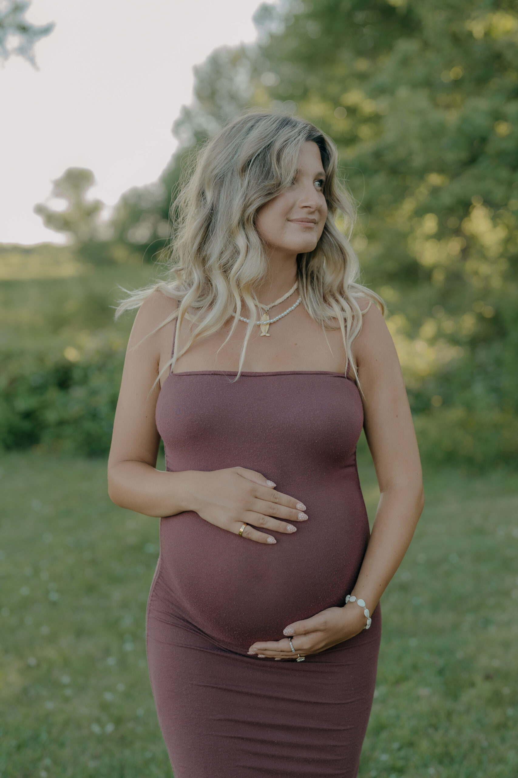 A pregnant wife stands facing forward, both hands resting on her belly as natural light highlights her expression through documentary wedding photography.