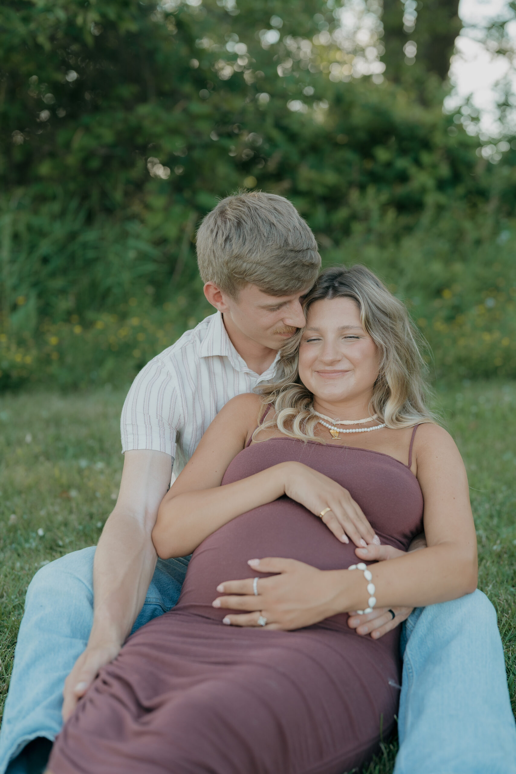 A husband sits behind his pregnant wife on the grass, resting his head near hers as documentary wedding photography captures a quiet, intimate moment.