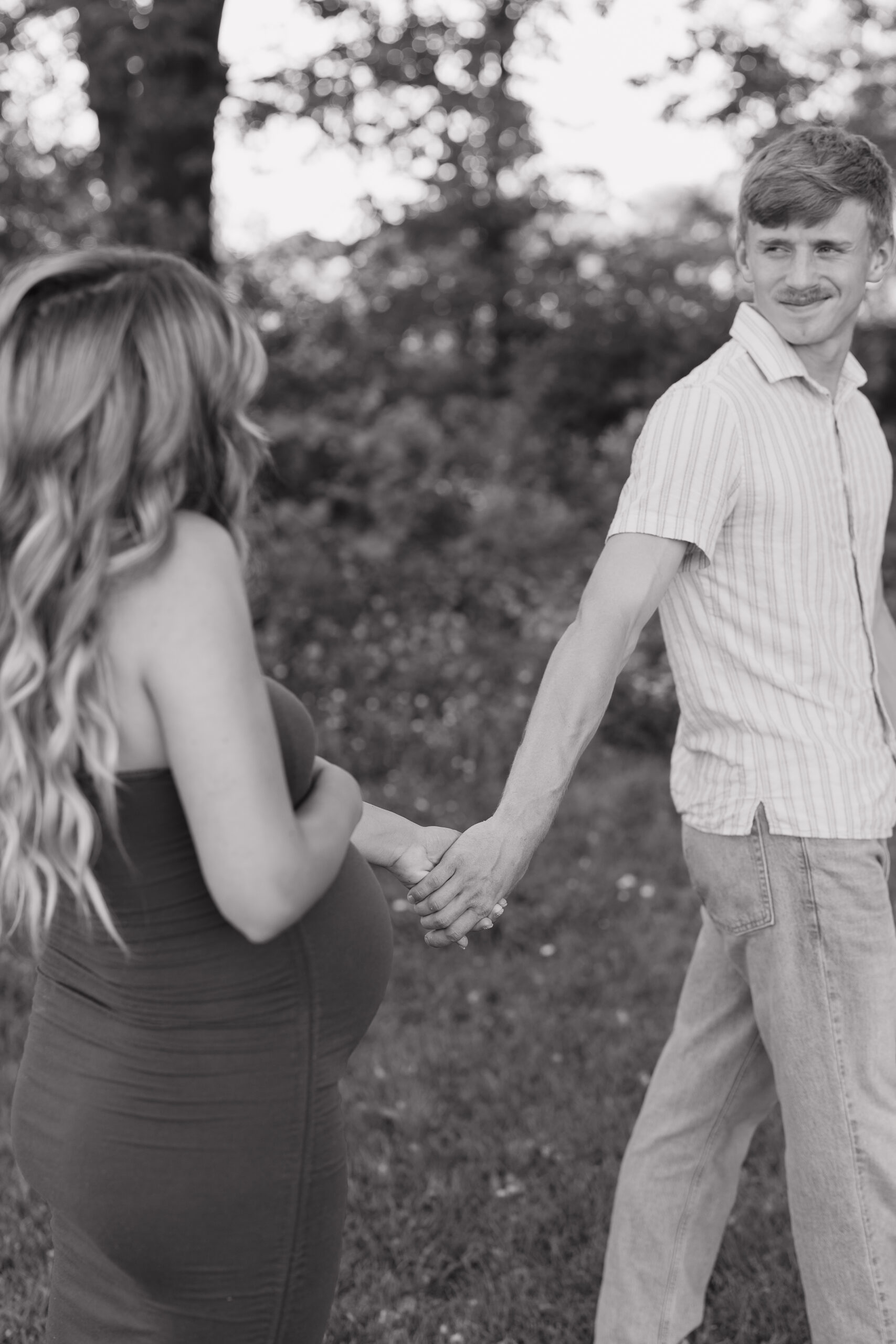 A husband gently holds his pregnant wife’s hand as they walk together through a grassy field, captured in black and white with the storytelling approach of documentary wedding photography.