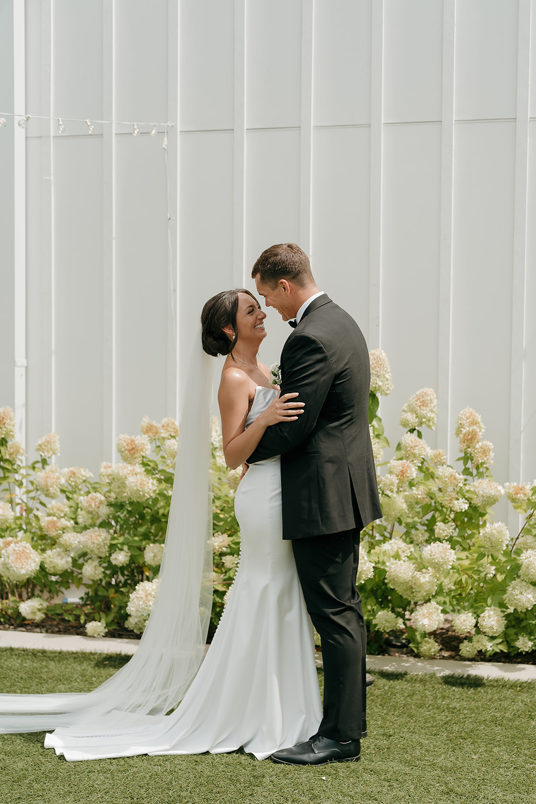 Full length portrait of the bride and groom standing together in front of hydrangeas and a white exterior wall at a Des Moines wedding venue
