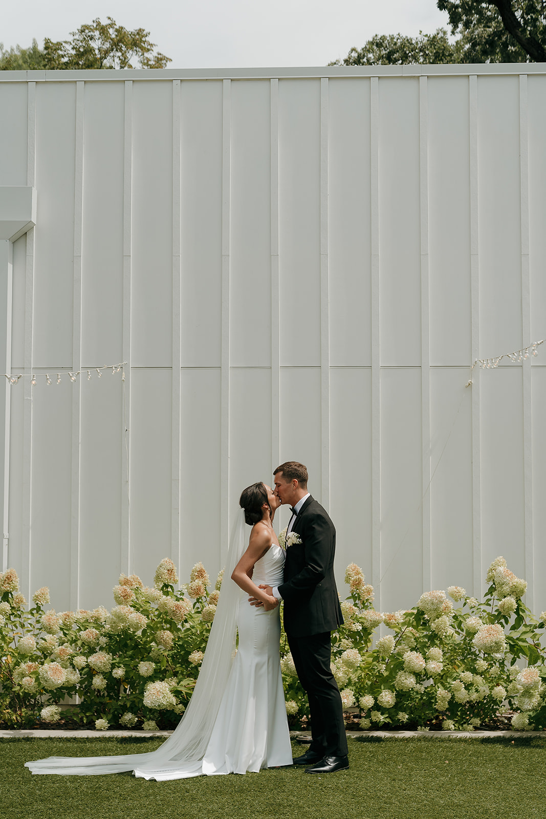Full length portrait of the bride and groom kissing outside a modern Des Moines wedding venue with floral landscaping.