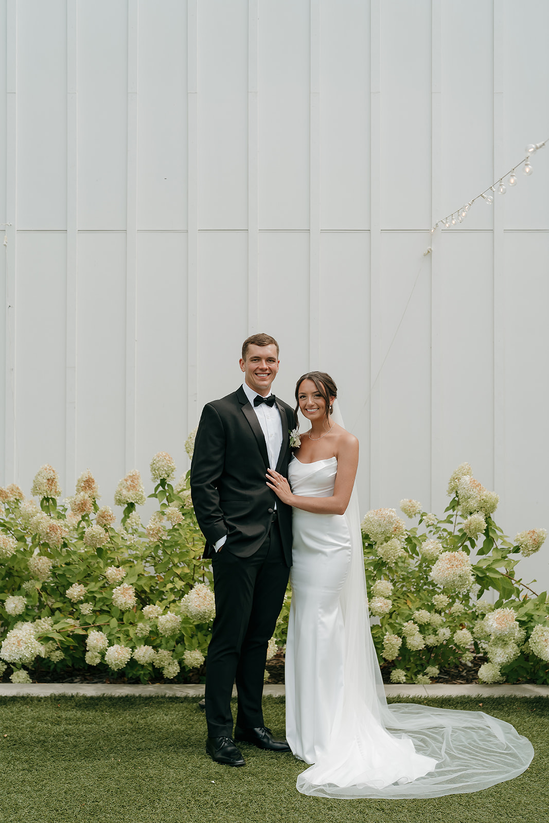 Full length portrait of the bride and groom standing together outside a modern Des Moines wedding venue with floral landscaping.