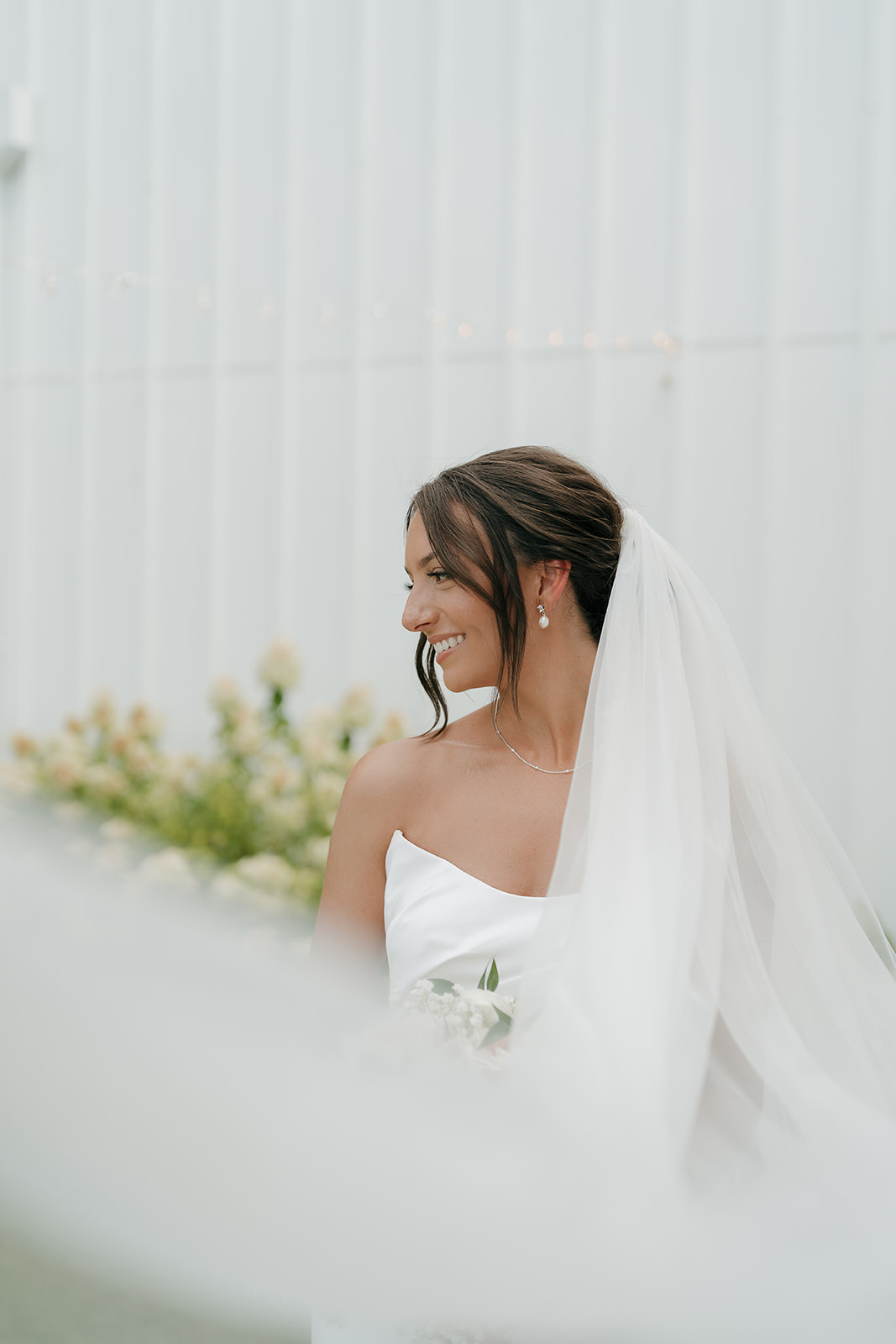 A bridal photo of the bride smiling and looking to the side while holding her bridal bouquet outside a modern Des Moines wedding venue with a white wall behind her.