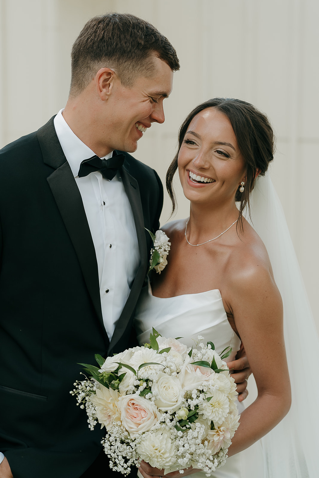 A portrait of the bride smiling and the groom smiling down at her during their couple portraits outside a modern Des Moines wedding venue with a white wall behind them