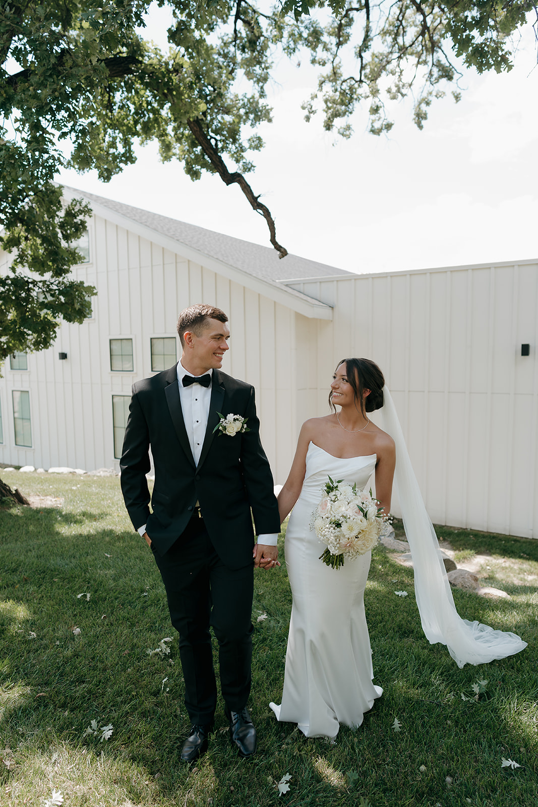 Bride and groom walking hand in hand outside near a white modern building, smiling at each other in natural daylight.
