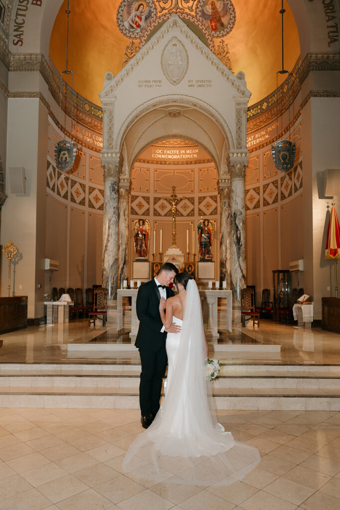 Bride and groom standing together at the altar inside an ornate Catholic church at a Des Moines wedding venue, surrounded by warm golden architecture and marble columns.