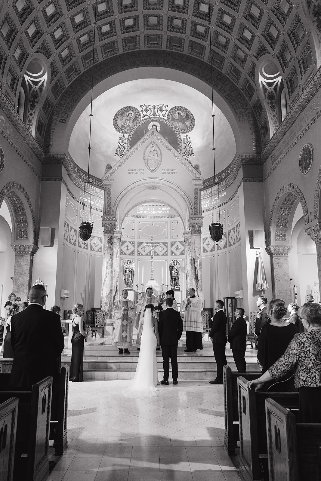 A black and white photo of the bride and groom standing together at the altar inside an ornate Catholic church at a Des Moines wedding venue, surrounded by warm golden architecture and marble columns.