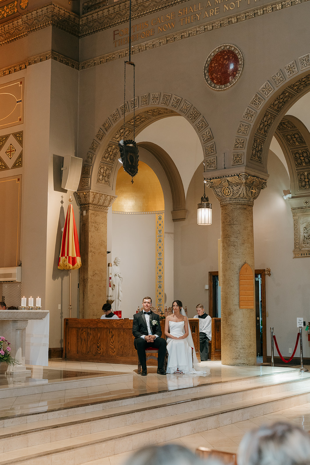 Bride and groom sitting together at the altar inside an ornate Catholic church at a Des Moines wedding venue, surrounded by warm golden architecture and marble columns.