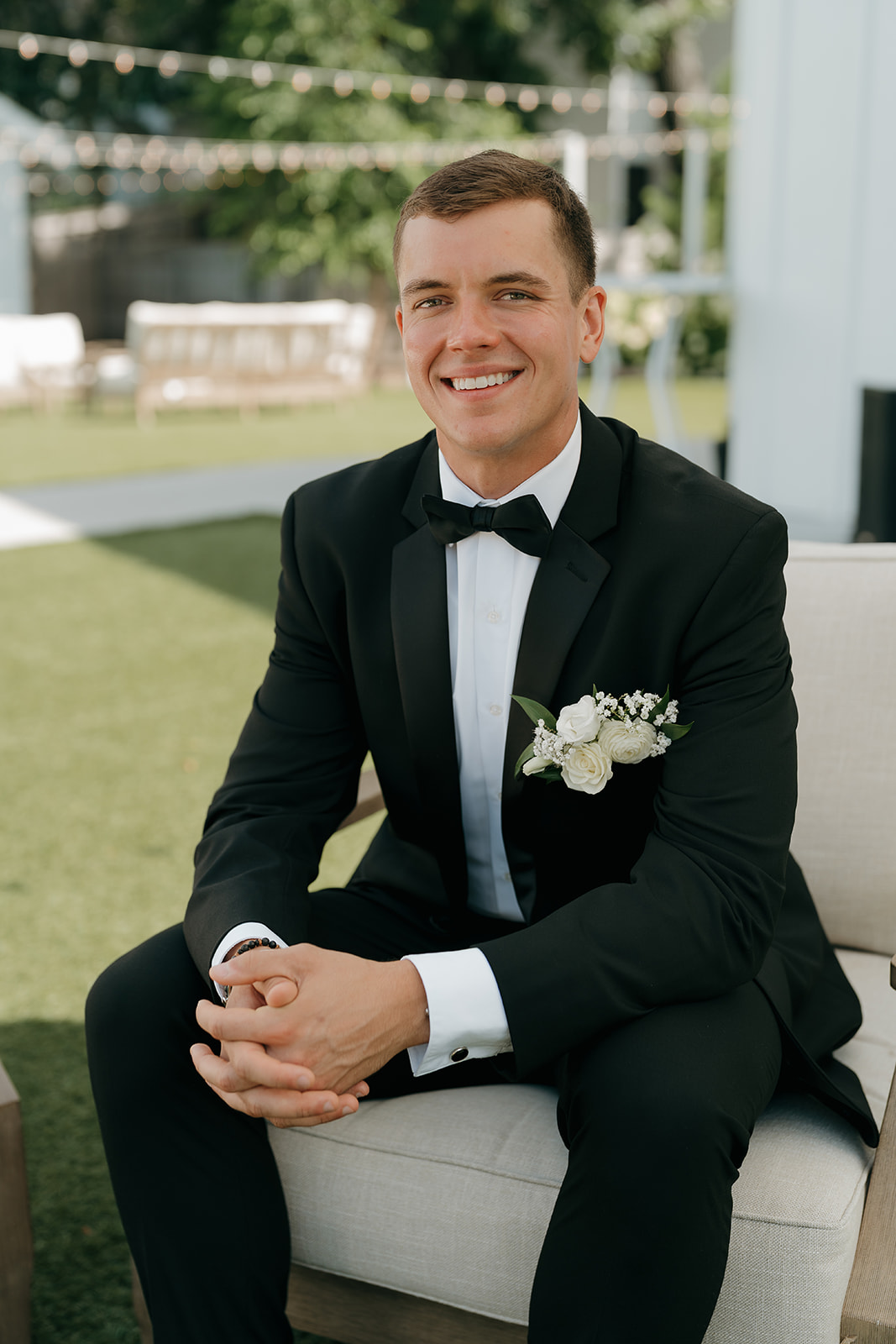 Groom sitting outdoors in a tuxedo with boutonnière, relaxed and smiling at this elegant Des Moines wedding venue during cocktail hour.