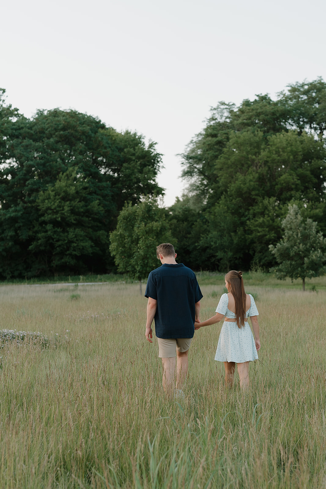 Couple walking hand in hand through a tall grassy field at sunset, surrounded by trees and wildflowers during a romantic engagement session.