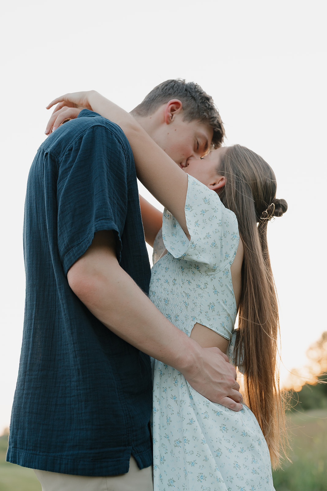 Golden hour kiss in an open field, soft light wrapping around the engaged couple