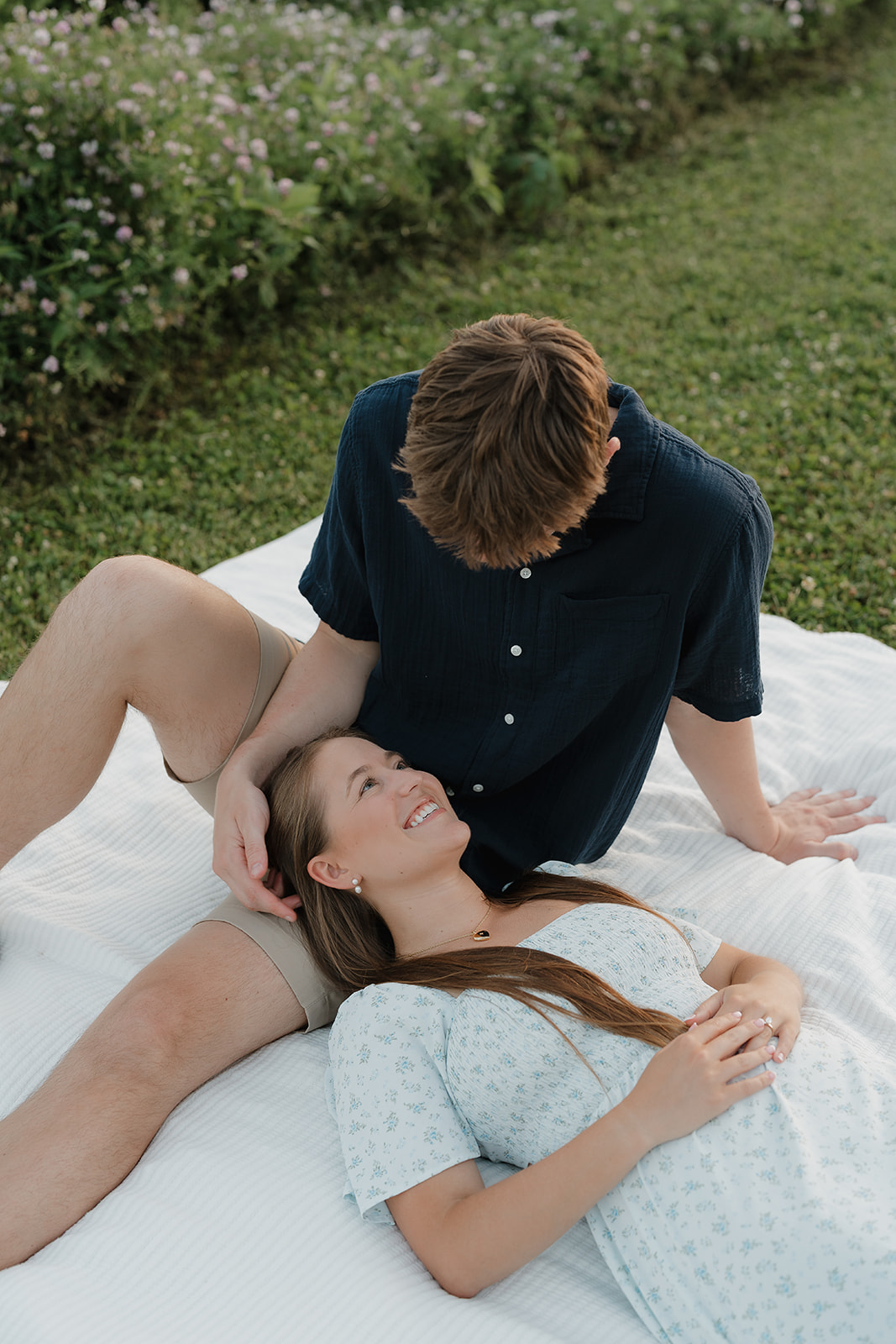 A couple sitting on a blanket in the grass while the girl rests her head on her fiances lap as they look at each other.