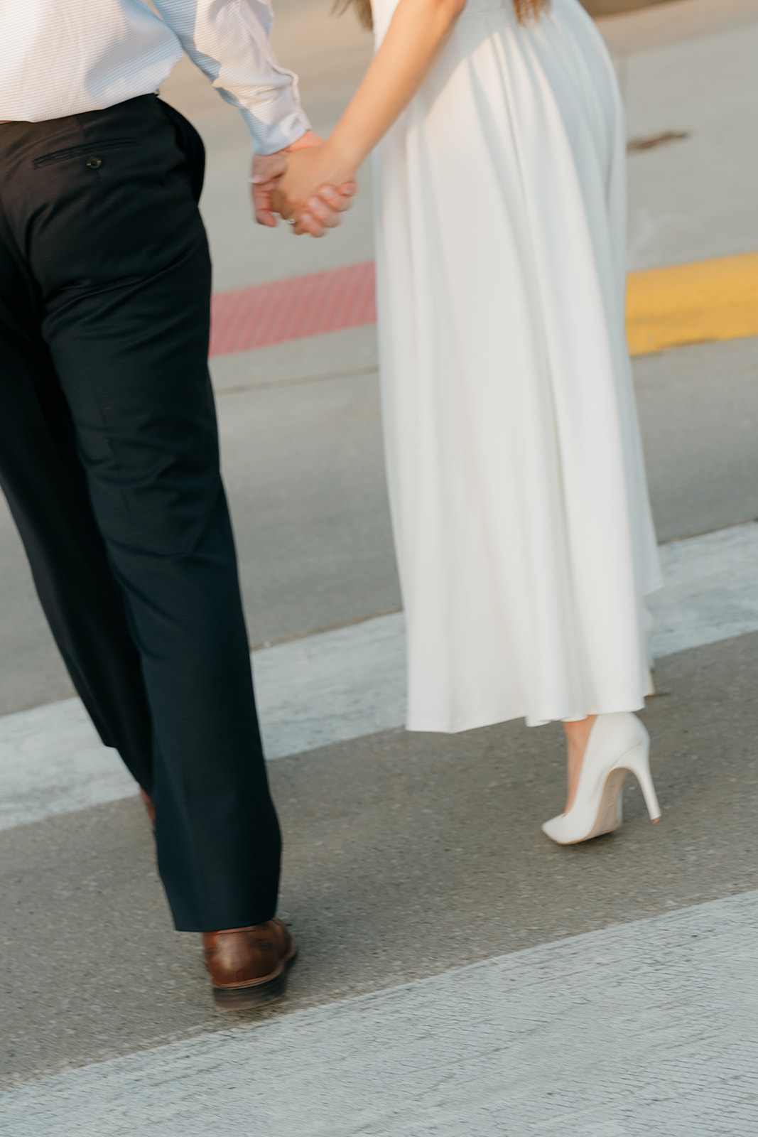 Close-up of a couple holding hands while walking across a city street, highlighting their formal engagement outfits and coordinated neutral tones.