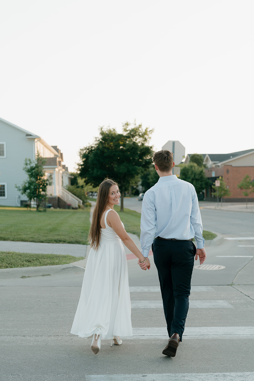 Couple holding hands while walking across a quiet city street during golden hour, dressed in classic neutral engagement outfits.