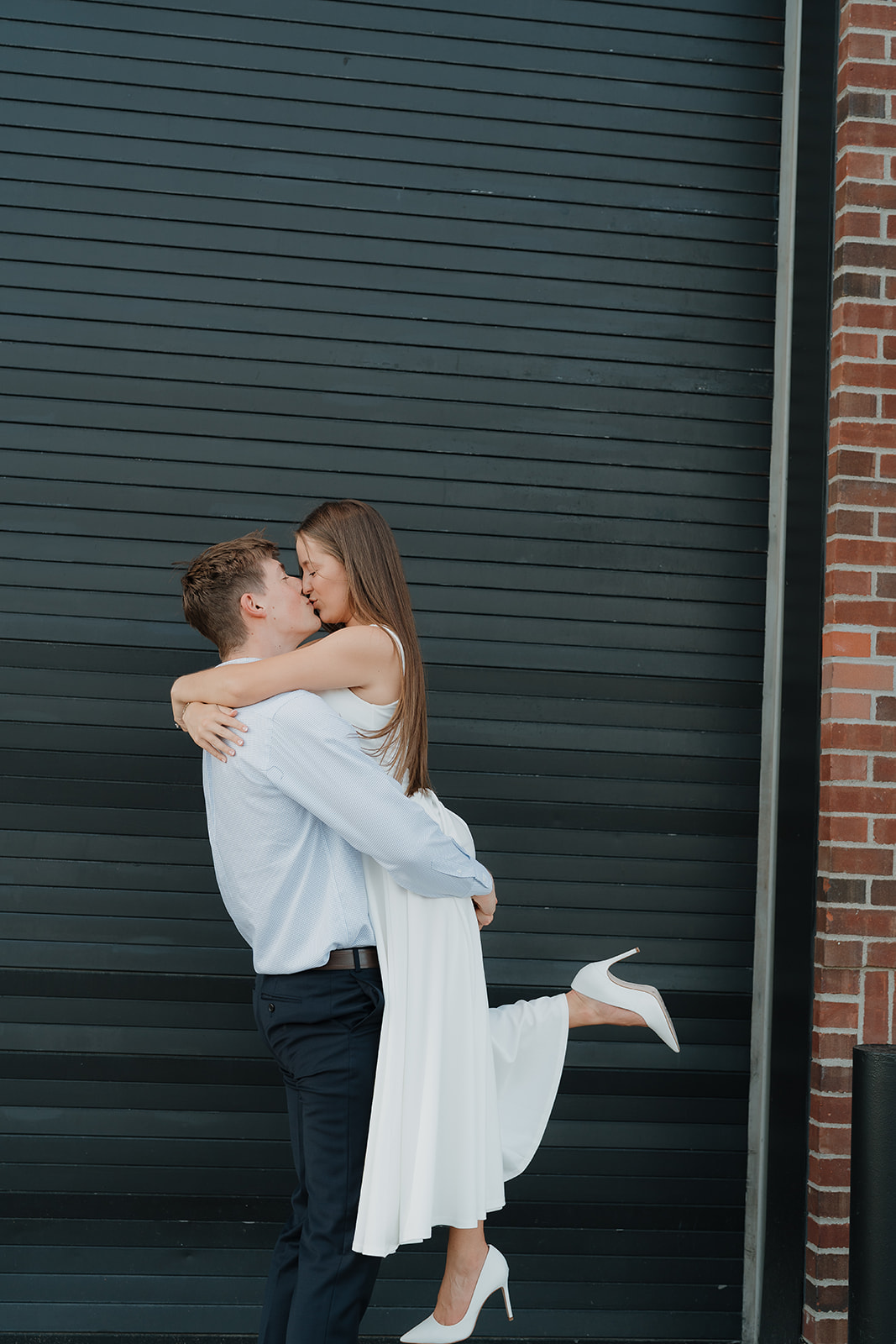 Couple kissing in front of a modern black industrial garage door, with the groom lifting the bride in a playful, candid engagement moment.