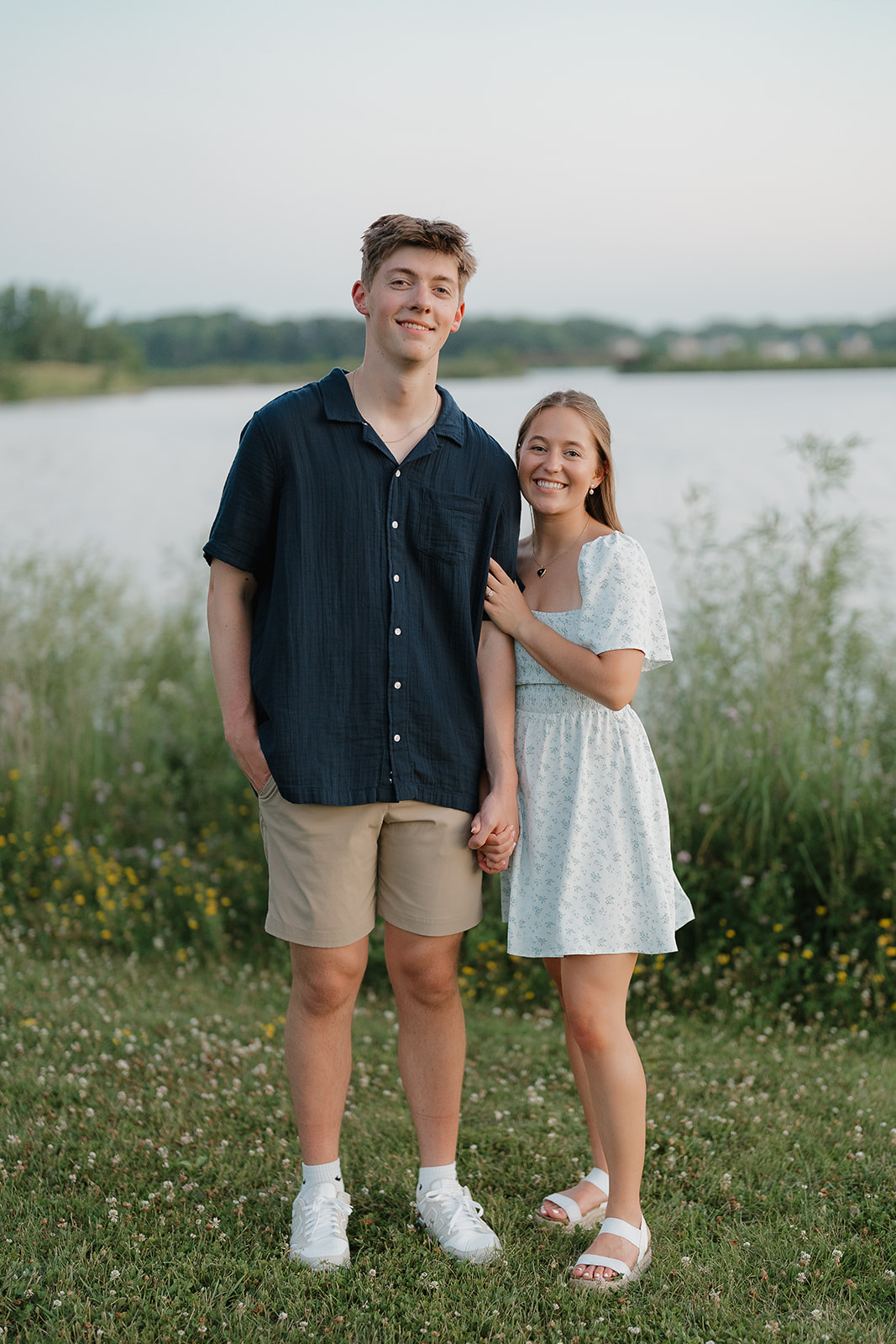 Couple standing together by the water in a grassy field, casual summer engagement photos
