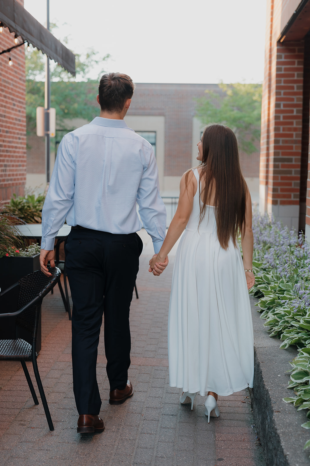 Couple walking hand in hand, the girl wearing a white midi dress and heels with the guy wearing dress pants and button up for a romantic engagement session