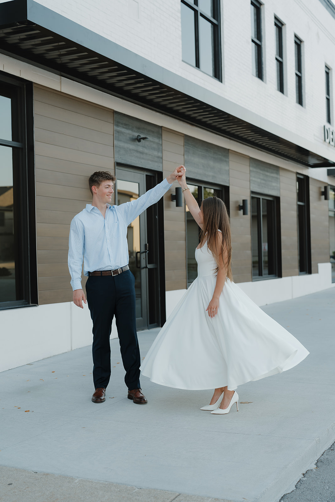 Playful twirling moment as the groom lifts his arm and spins his bride-to-be in front of a modern storefront with clean architectural lines.