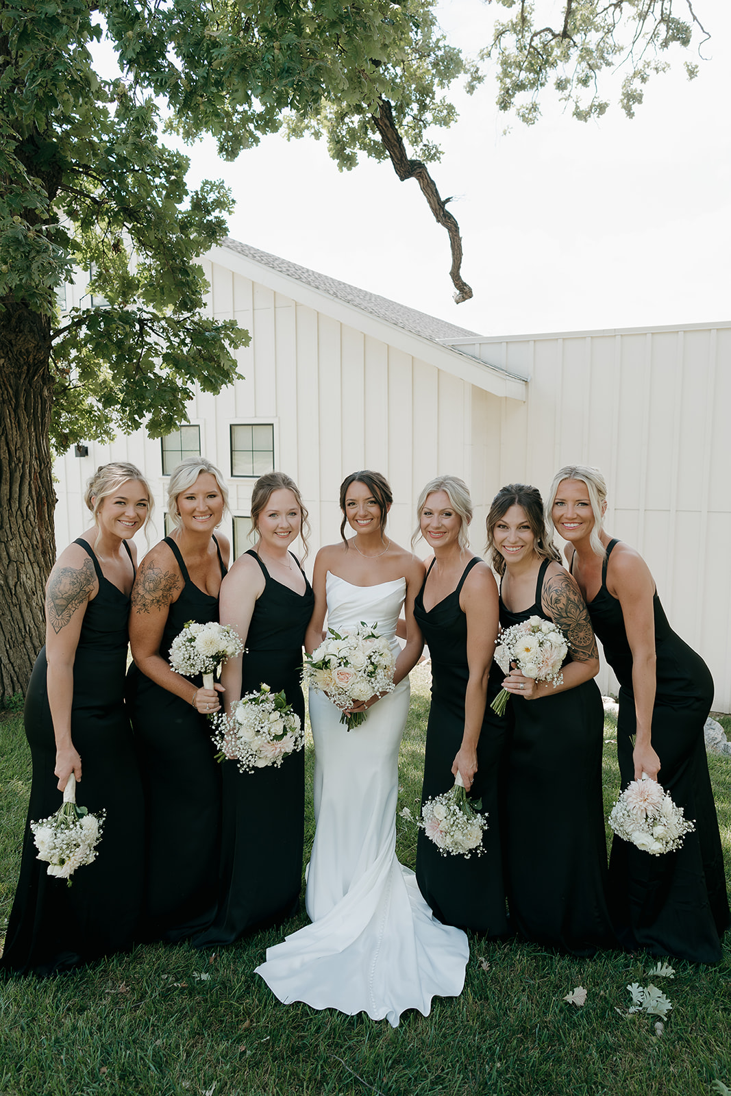 A bride in a sleek white gown stands with six bridesmaids in black dresses, all holding white floral bouquets outside a modern white barn venue — a striking choice of wedding colors for a summer wedding.