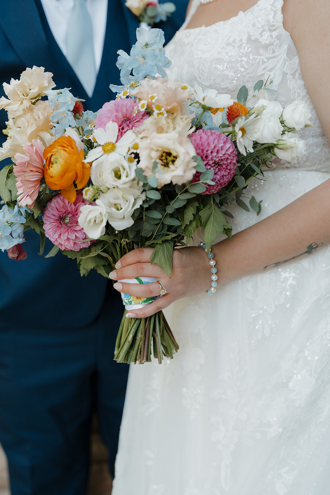 A close-up of a bride holding a lush bouquet of orange ranunculus, pink dahlias, blue delphinium, and white daisies — a vibrant and joyful choice of wedding colors for a summer wedding.