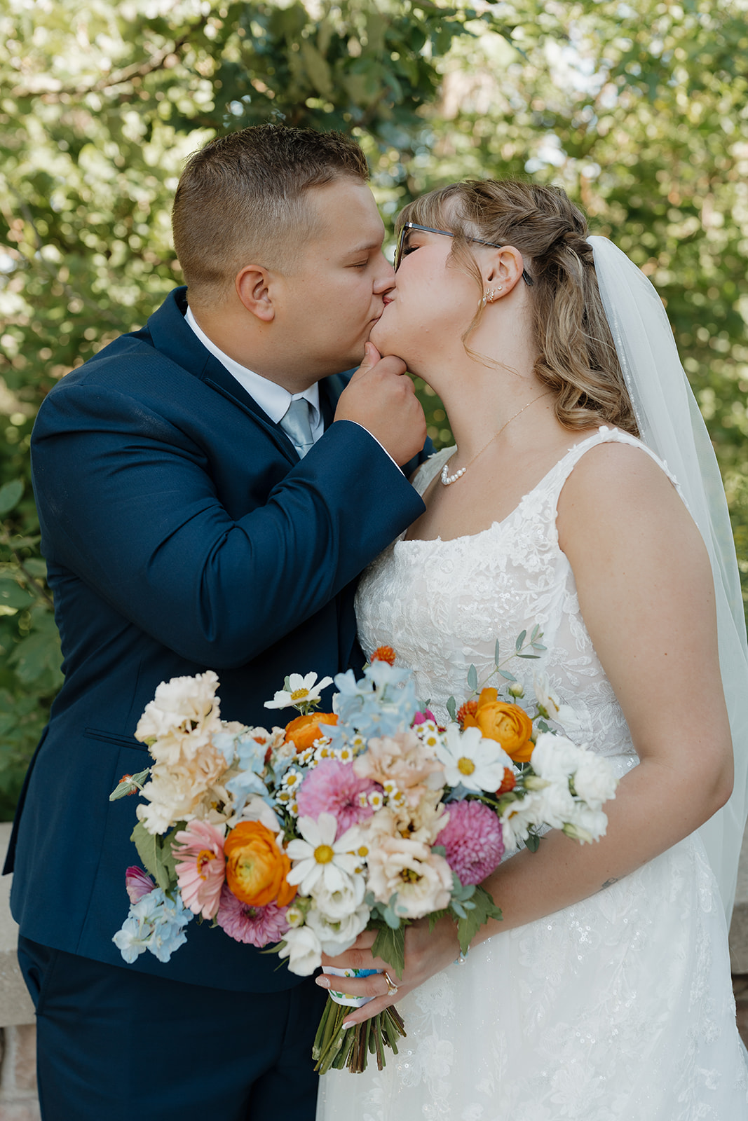 A groom in a navy suit kisses his bride while she holds a vibrant bouquet of orange ranunculus, pink dahlias, blue delphinium, and white daisies — a bright and joyful choice of wedding colors for a summer wedding.
