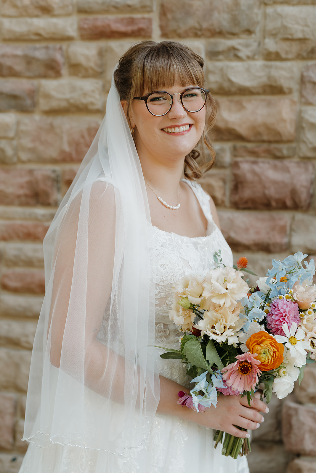 A smiling bride in a lace gown and veil holds a vibrant bouquet of peach, blue, orange, and pink blooms in front of a stone wall.