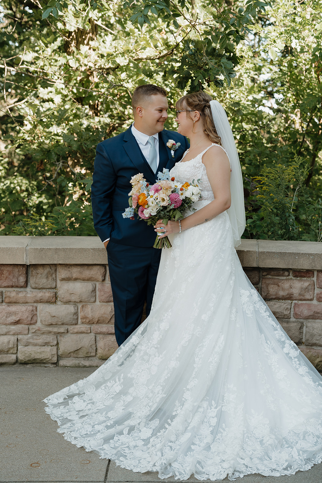 A bride and groom smile at each other outside against a stone wall, her lace cathedral-train gown and colorful bouquet of pink, orange, and blue blooms showing off beautiful wedding colors for a summer wedding.