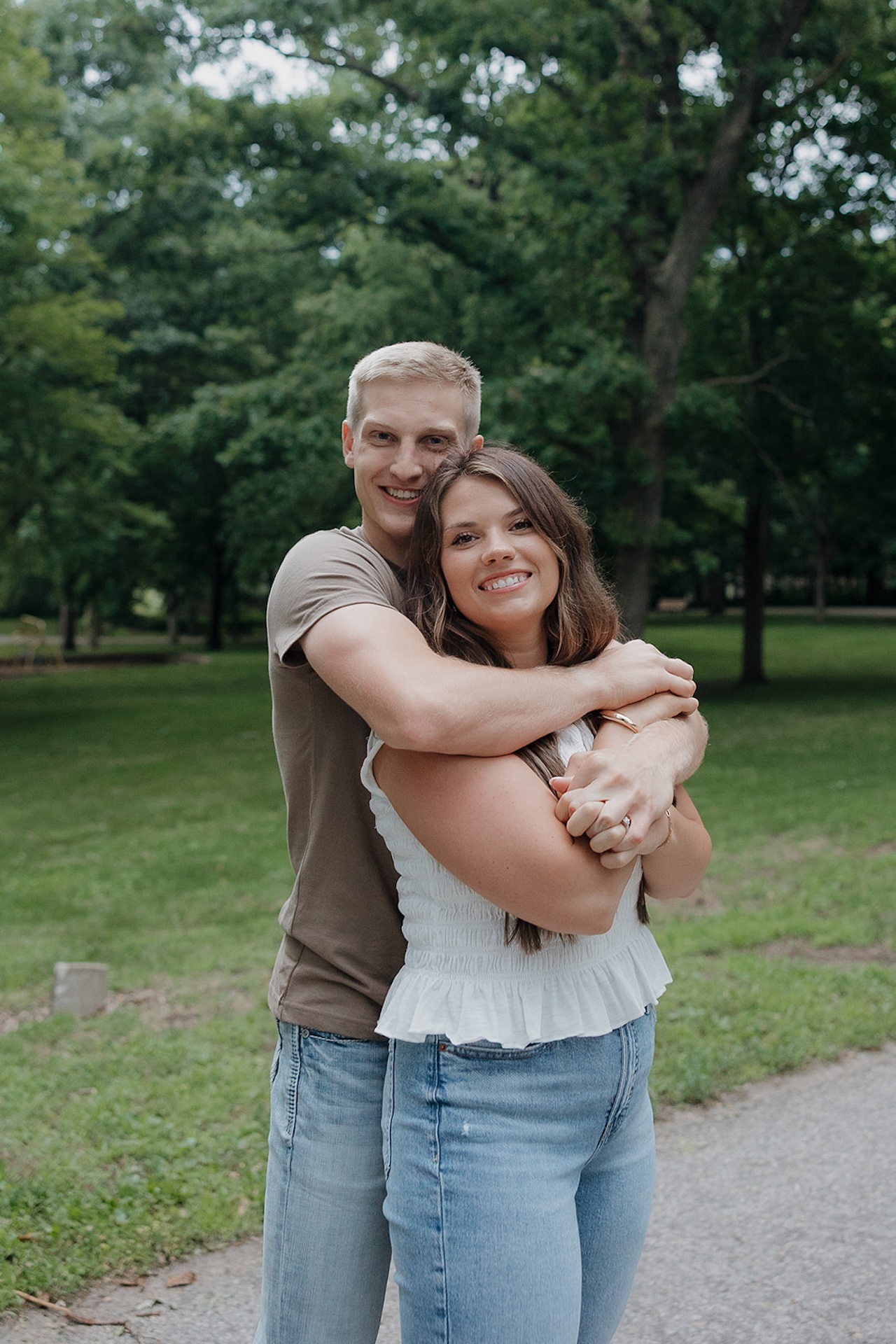 A smiling engaged couple hugging together on a park path surrounded by lush green trees in Iowa