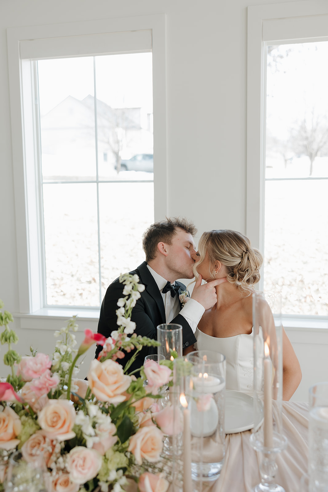A bride and groom share a kiss at their sweetheart table, framed by bright windows and lush pink and peach floral centerpieces — a beautiful example of soft wedding colors for a summer wedding.