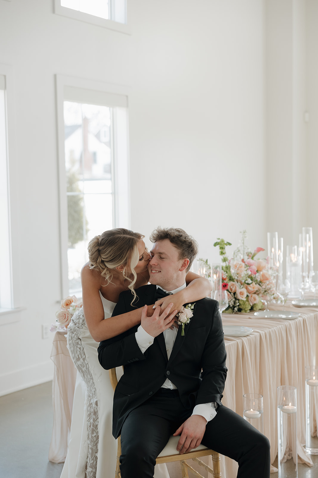 A bride kisses her groom on the cheek as he smiles at their sweetheart table, surrounded by candlelight and a blush floral arrangement in a bright white venue.