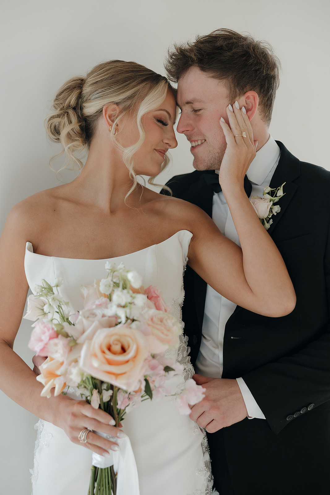 A bride and groom lean foreheads together against a white wall, her holding a soft peach and blush rose bouquet as he smiles warmly in a black tuxedo.
